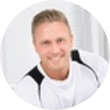 Lächelnde Person in einem weißen Hemd mit schwarzen Akzenten, die vor einem hellen Hintergrund im Innenbereich sitzt und die Webdesign Agentur Buchholz in der Nordheide repräsentiert.