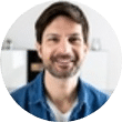 Ein lächelnder Mann mit dunklem Haar und Bart, der ein blaues Hemd trägt, drinnen vor unscharfem Hintergrund, im Büro einer Webdesign-Agentur in Buchholz in der Nordheide.
